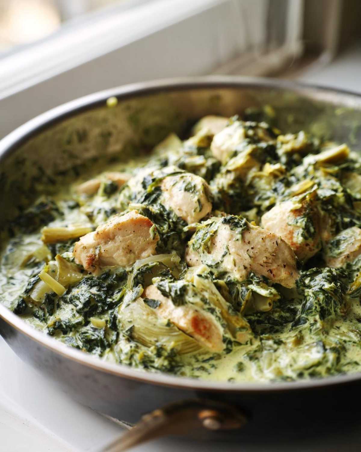Close-up of chunks of chicken breast mixed into a rich, creamy spinach artichoke sauce in a skillet.