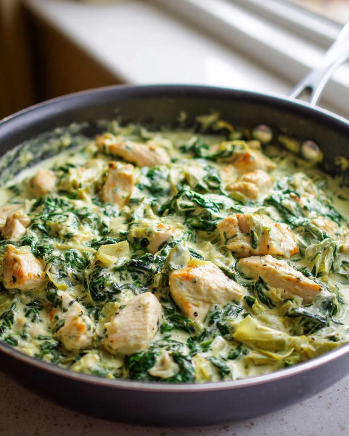 Close-up of Creamy Spinach Artichoke Chicken Skillet cooking in a dark skillet with chunks of chicken and green spinach.