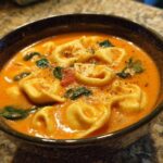A close-up of a dark bowl filled with rich, orange Creamy Tomato Tortellini Soup, featuring tortellini pasta, spinach, and grated cheese.