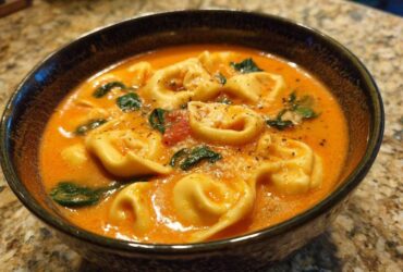 A close-up of a dark bowl filled with rich, orange Creamy Tomato Tortellini Soup, featuring tortellini pasta, spinach, and grated cheese.