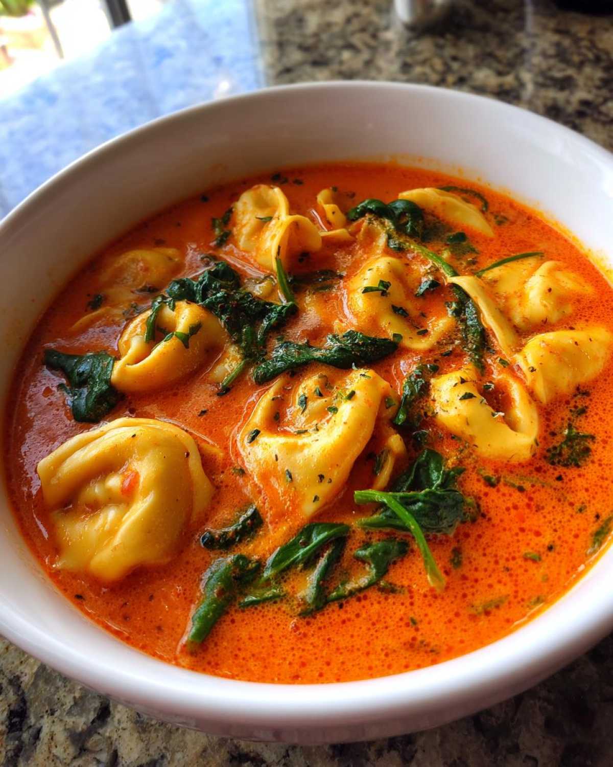 Close-up of a white bowl filled with vibrant Creamy Tomato Tortellini Soup, featuring cheese tortellini and wilted spinach.
