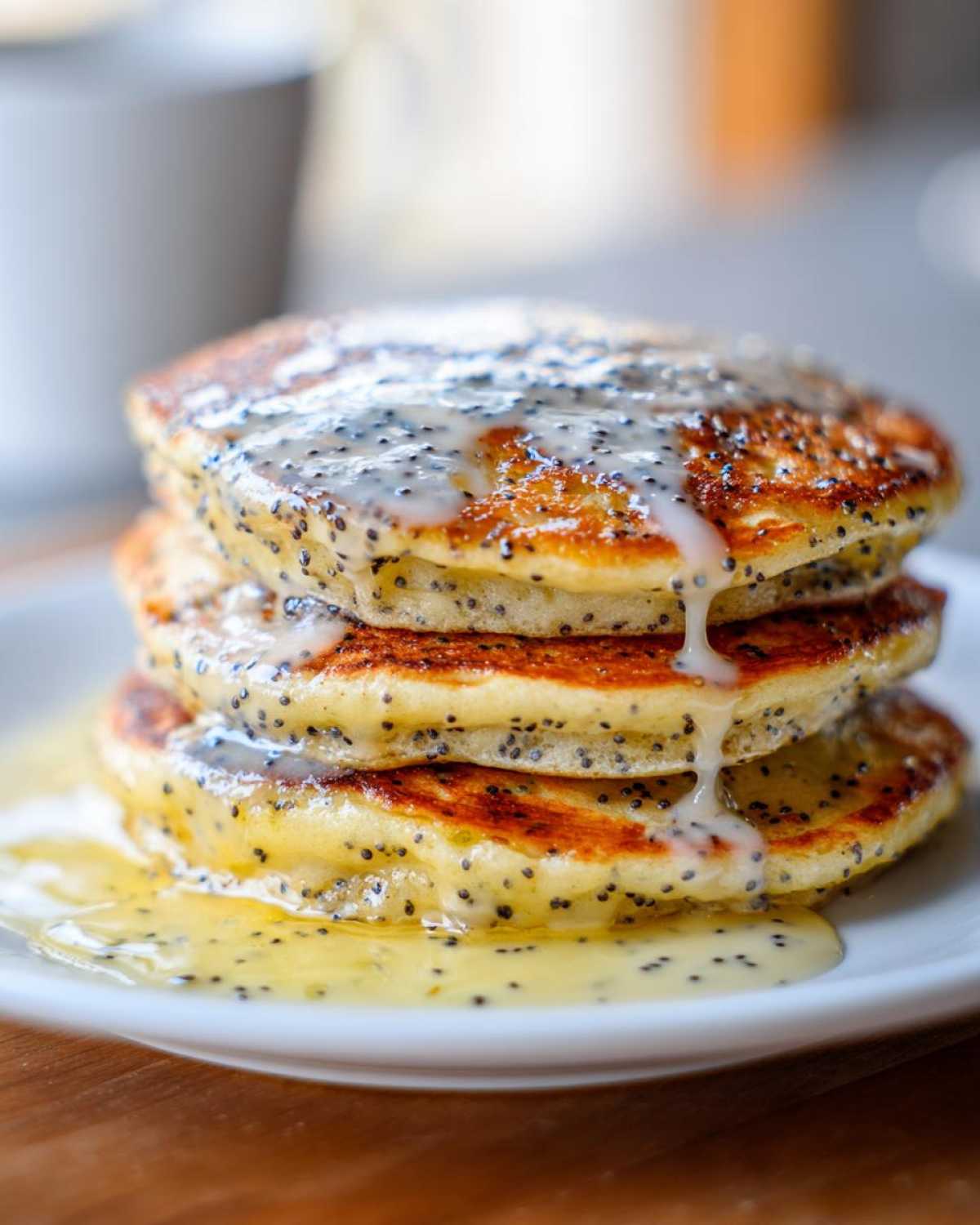 A stack of three fluffy Vegan Lemon Poppy Seed Pancakes drizzled with lemon glaze and poppy seeds.