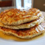 A stack of three fluffy Vegan Lemon Poppy Seed Pancakes drizzled with syrup.