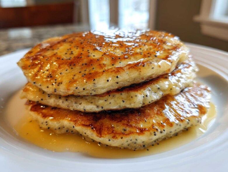 A stack of three fluffy Vegan Lemon Poppy Seed Pancakes drizzled with syrup.