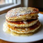 A stack of three fluffy Vegan Lemon Poppy Seed Pancakes drizzled with lemon glaze and poppy seeds.