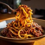 A fork lifts a generous twirl of thick pasta coated in rich Vegan Lentil Bolognese sauce.