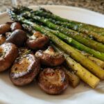 Roasted asparagus spears and browned mushrooms seasoned with herbs on a white plate.