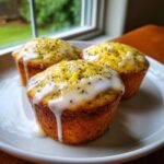 Three bright yellow Glazed Lemon Poppy Seed Muffins topped with thick white icing and poppy seeds, sitting on a white plate.