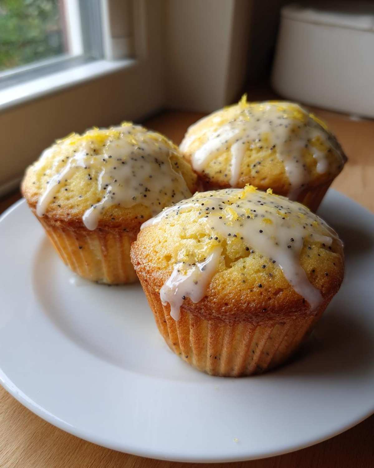 Three freshly baked Glazed Lemon Poppy Seed Muffins topped with white icing and lemon zest, sitting on a white plate.