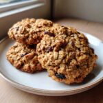 Three freshly baked Gluten-Free Oatmeal Raisin Cookies stacked slightly on a light ceramic plate near a window.