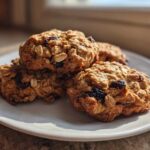 A stack of four golden-brown Gluten-Free Oatmeal Raisin Cookies, studded with visible oats and dark raisins, resting on a white plate.