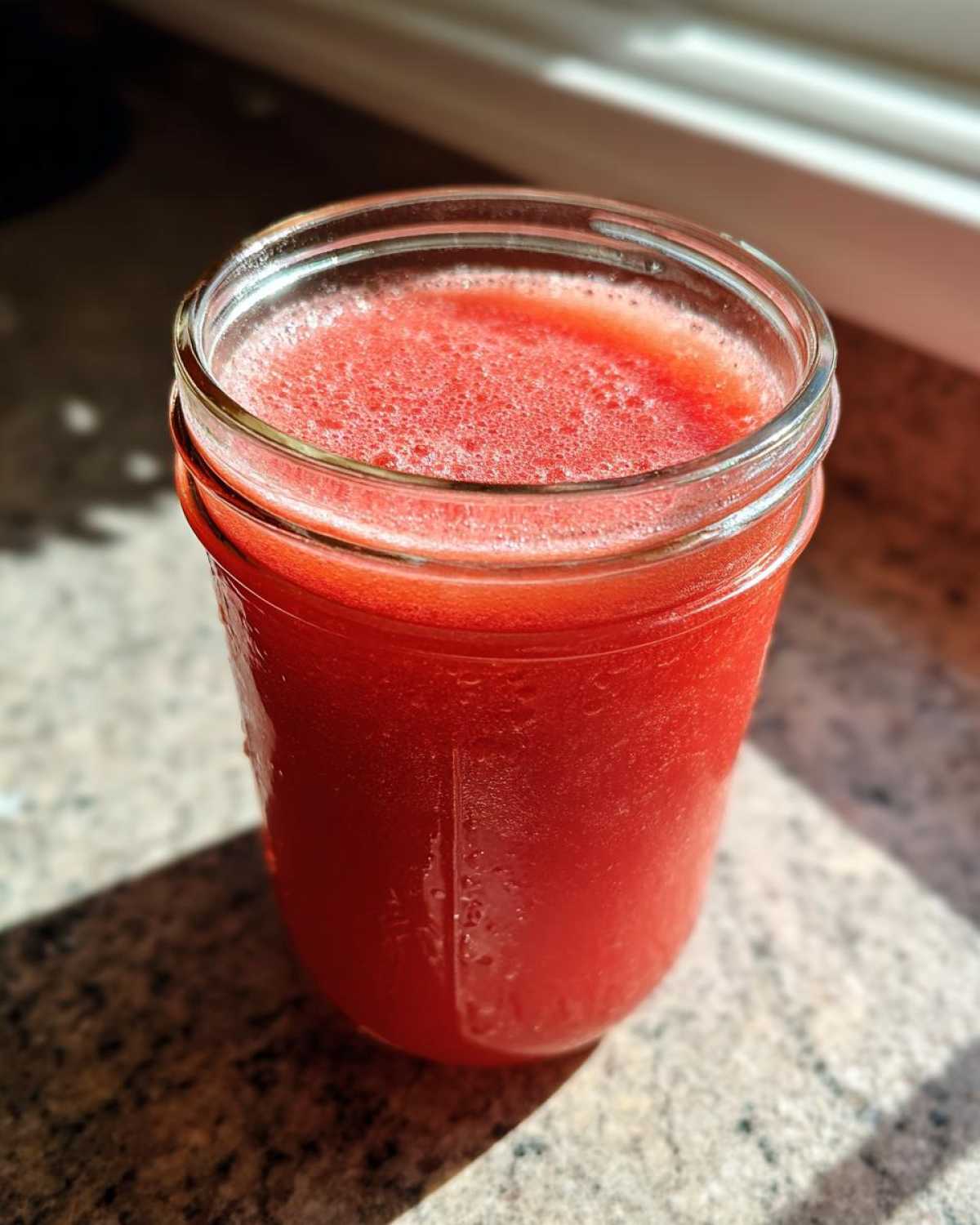 A glass jar filled with bright red liquid, likely Homemade Strawberry Lemonade Concentrate, sitting on a speckled countertop in sunlight.