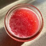 Overhead view of bright red liquid with foam, likely Homemade Strawberry Lemonade Concentrate, in a glass jar.