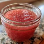 Close-up of bright red Homemade Strawberry Lemonade Concentrate stored in a small glass jar.