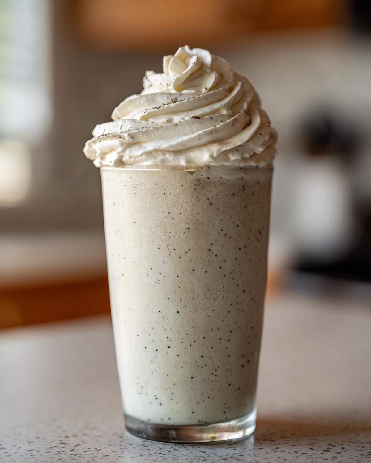 A tall glass filled with a creamy Homemade Vanilla Bean Frappuccino, topped with a swirl of whipped cream and vanilla bean specks.