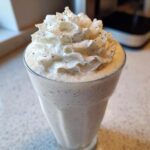 Close-up of a tall glass filled with Homemade Vanilla Bean Frappuccino, topped generously with whipped cream and vanilla bean specks.