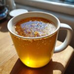 Close-up of warm Honey Lemon Ginger Tea in a white mug, showing floating spices on the surface.