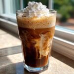 A tall glass of Iced Mocha showing swirls of cream mixing with dark coffee, topped with whipped cream.