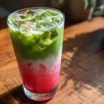 A tall glass showing the distinct layers of an Iced Strawberry Matcha Latte: bright red strawberry base, white milk, and green matcha.