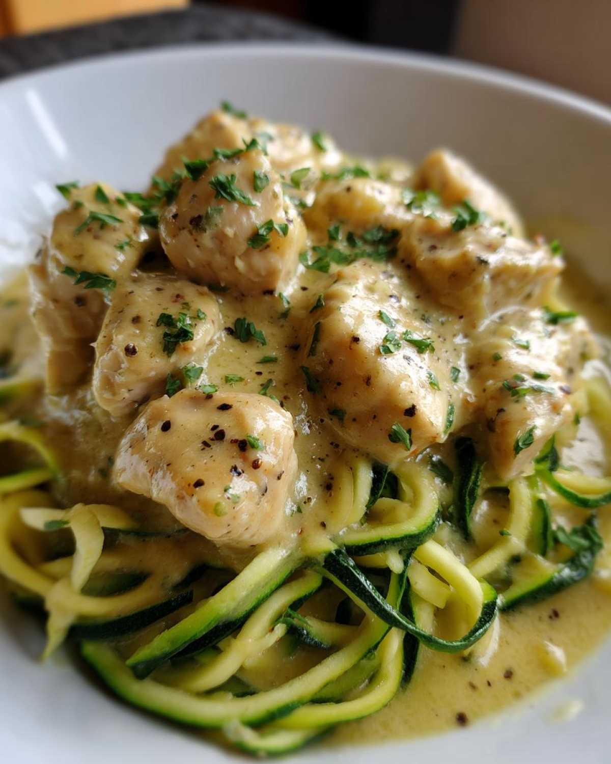 A close-up of Keto Chicken Alfredo featuring tender chicken pieces in a creamy sauce over bright green zucchini noodles, garnished with parsley.