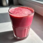 A close-up of a vibrant, thick, pink-red Keto Strawberry Cheesecake Smoothie in a clear glass, sitting in sunlight.