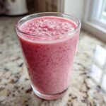A close-up of a thick, pink Keto Strawberry Cheesecake Smoothie served in a clear glass on a granite countertop.
