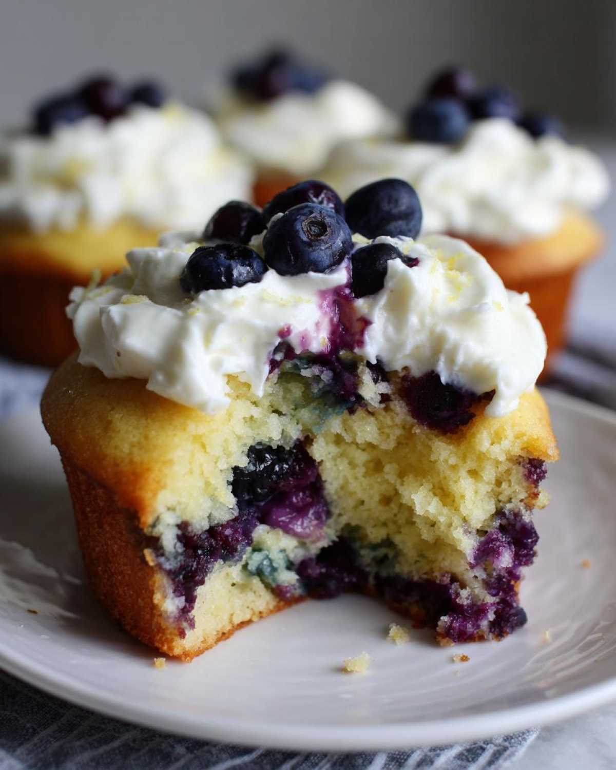 Close-up of a Lemon Blueberry Cupcake with cream cheese frosting, showing a bite taken out revealing the moist interior.