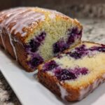 A moist Lemon Blueberry Yogurt Loaf, partially sliced, showing juicy blueberries and a sweet lemon glaze.