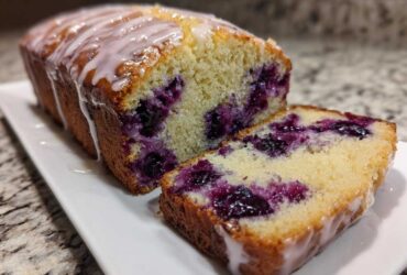 A moist Lemon Blueberry Yogurt Loaf, partially sliced, showing juicy blueberries and a sweet lemon glaze.