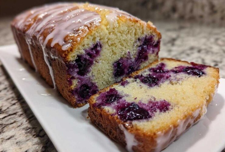 A moist Lemon Blueberry Yogurt Loaf, partially sliced, showing juicy blueberries and a sweet lemon glaze.
