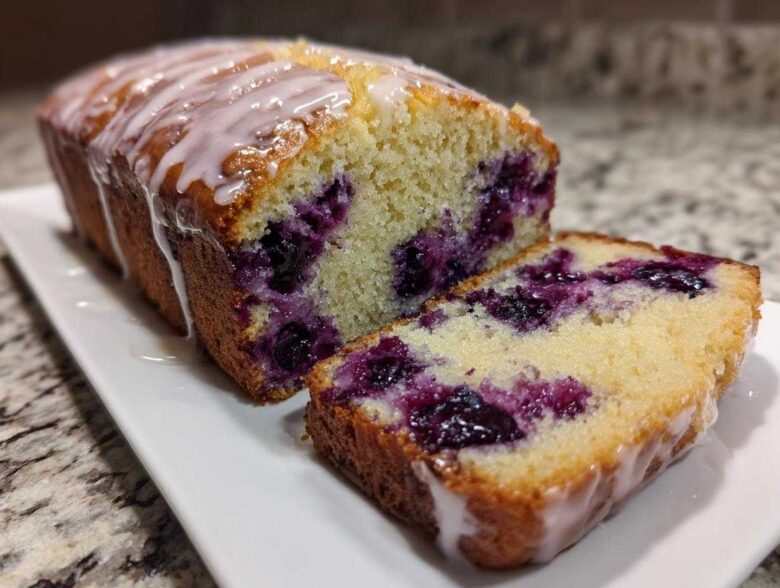 A moist Lemon Blueberry Yogurt Loaf, partially sliced, showing juicy blueberries and a sweet lemon glaze.
