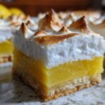A close-up, featured shot of one square of Lemon Meringue Pie Bars showing the crust, bright lemon filling, and toasted meringue topping.