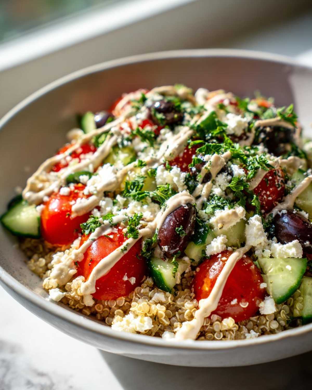 Close-up of a vibrant Mediterranean Quinoa Bowl topped with tomatoes, cucumbers, olives, feta, and drizzled with lemon tahini sauce.