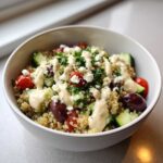 A close-up of a Mediterranean Quinoa Bowl topped with feta, cucumbers, tomatoes, olives, and drizzled with lemon tahini sauce.