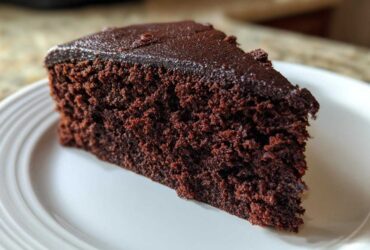 Close-up of a rich, dark slice of Gluten-Free Chocolate Cake with a thin chocolate glaze on a white plate.