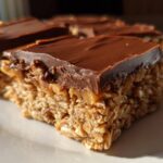 A close-up, sunlit shot of a square of No-Bake Chocolate Peanut Butter Oat Bars showing the thick oat base and chocolate topping.