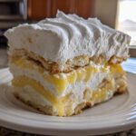 A tall slice of No-Bake Lemon Icebox Cake showing layers of lemon filling, whipped cream, and vanilla wafers.
