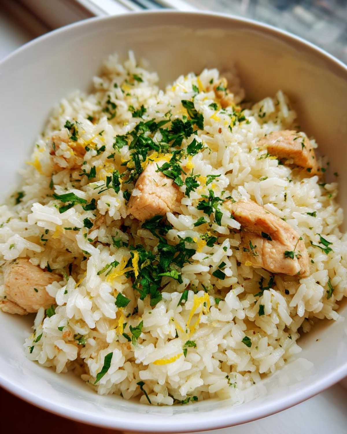 Close-up of fluffy white rice mixed with chunks of chicken, topped with fresh parsley and lemon zest in One-Pot Chicken and Rice with Lemon and Herbs.