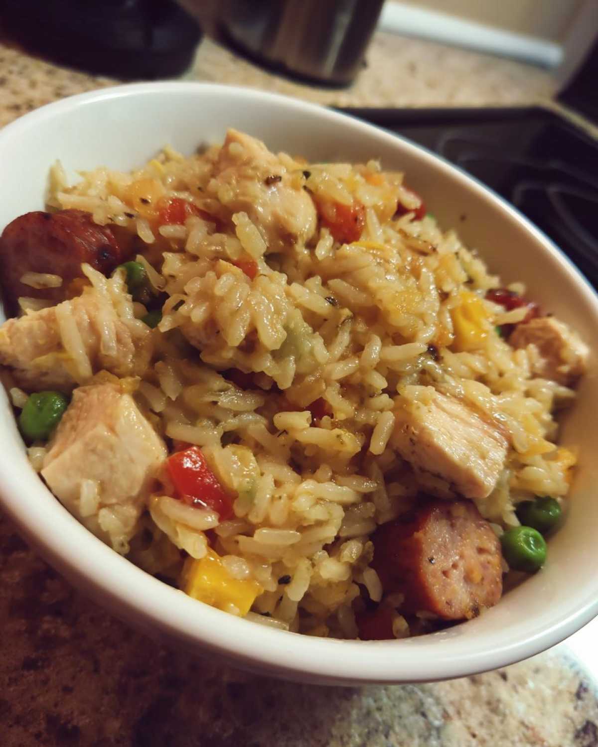 Close-up of a white bowl filled with moist One-Pot Chicken Jambalaya, featuring rice, chicken chunks, sausage slices, and peas.