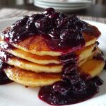 A stack of golden pancakes generously topped with rich, dark Homemade Blueberry Syrup and whole berries.