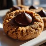 A close-up, sunlit photo of a freshly baked Peanut Butter Blossom Cookie with a shiny chocolate kiss pressed into the center.
