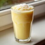 A tall glass filled with a thick, yellow Pineapple Coconut Smoothie, topped with shredded coconut.