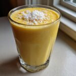 A tall glass filled with a thick, bright yellow Pineapple Coconut Smoothie, topped with shredded coconut.