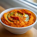 Close-up of vibrant orange Roasted Red Pepper Hummus drizzled with olive oil and herbs in a white bowl.