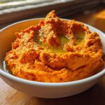 A close-up of vibrant orange Roasted Red Pepper Hummus drizzled with olive oil and herbs in a white bowl.