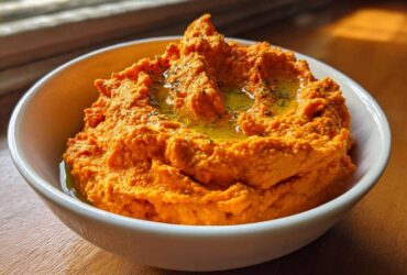 A close-up of vibrant orange Roasted Red Pepper Hummus drizzled with olive oil and herbs in a white bowl.