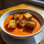 A white bowl filled with vibrant orange Roasted Tomato Basil Soup topped with five golden, crispy grilled cheese croutons.