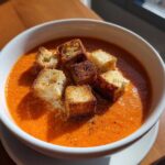 A white bowl filled with vibrant orange Roasted Tomato Basil Soup, topped with golden grilled cheese croutons and cracked black pepper.