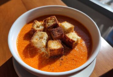 A white bowl filled with vibrant orange Roasted Tomato Basil Soup, topped with golden grilled cheese croutons and cracked black pepper.