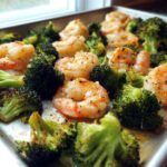 Close-up of Sheet Pan Garlic Butter Shrimp and Broccoli seasoned with pepper, glistening on a metal baking sheet.